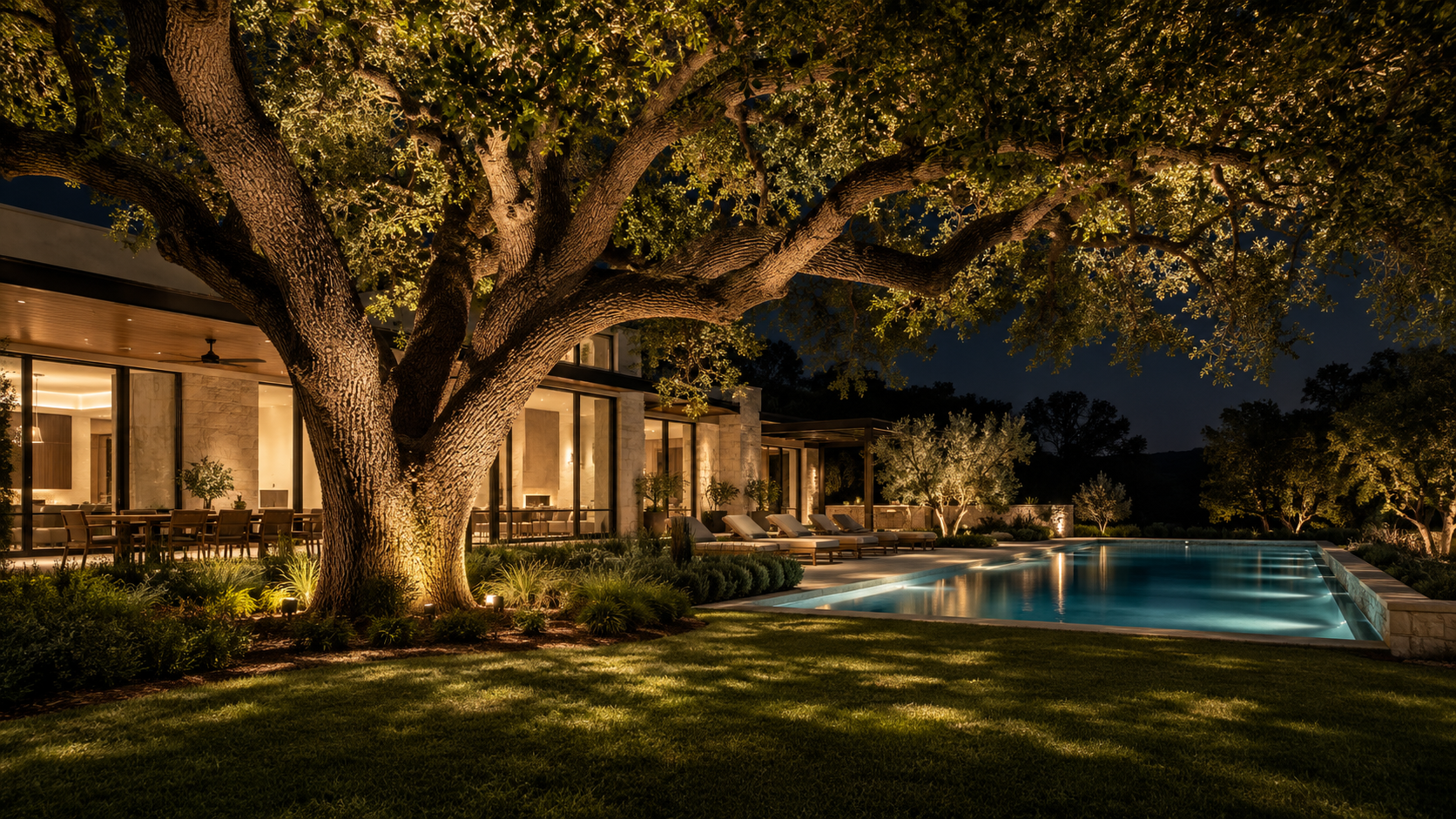 Mature live oak uplit at night above a lawn and pool behind a custom home